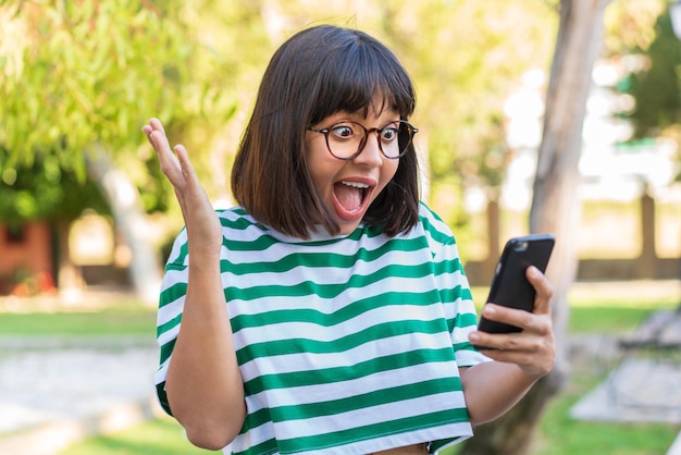 Young brunette woman in the park looking at the camera while using the mobile with surprised expression