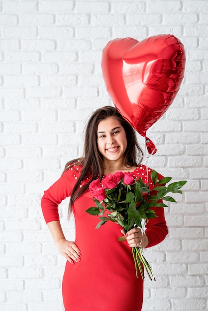 Young brunette woman in red dress holding a red heart balloon and flowers