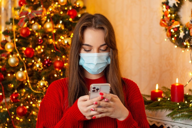 Young brunette woman in red sweater and protective medical mask standing at the background of Christmas tree chatting online Coronavirus distance communication winter holidays