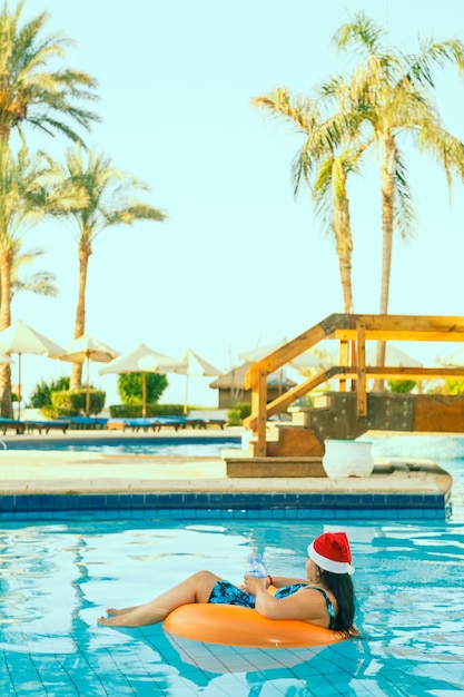 Young brunette woman in santa claus hat in a swimming circle in the pool during christmas holidays.