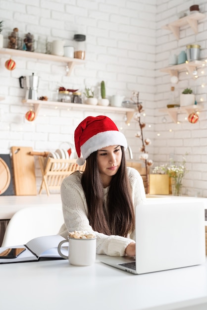 Young brunette woman in santa hat chatting with friends using her laptop at the kitchen