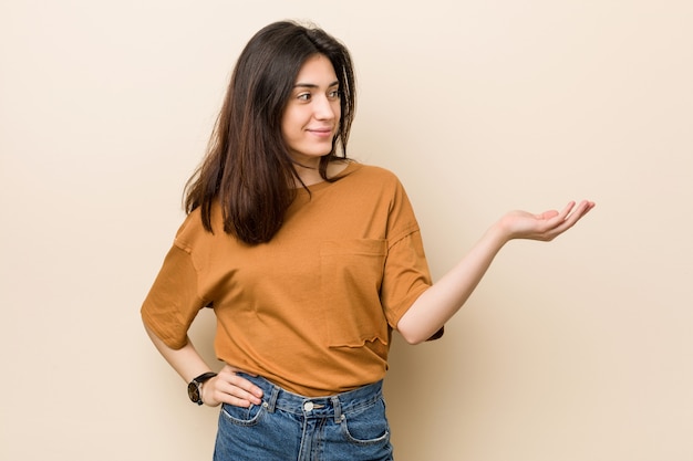 Young brunette woman showing a copy space on a palm and holding another hand on waist