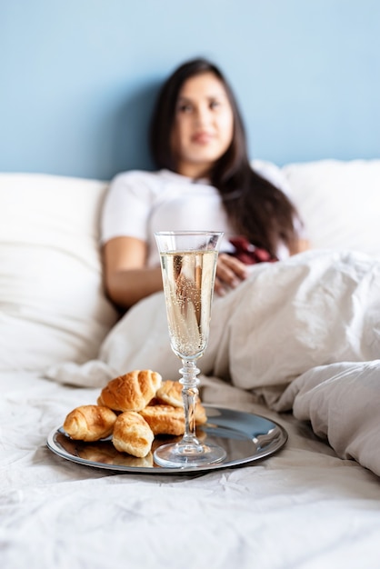 Young brunette woman sitting awake in the bed with red heart shaped balloons and decorations drinking champagne eating croissants