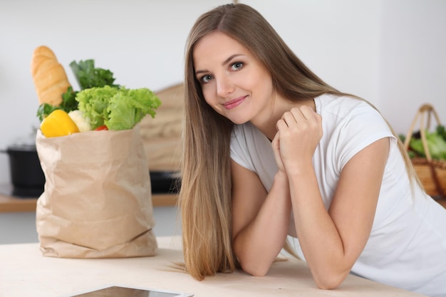 Young brunette woman smiling while sitting at the table near paper bag full of vegetables and fruits Concept of food buying and kitchen lifetime