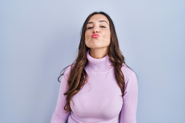 Young brunette woman standing over blue background looking at the camera blowing a kiss on air being lovely and sexy love expression