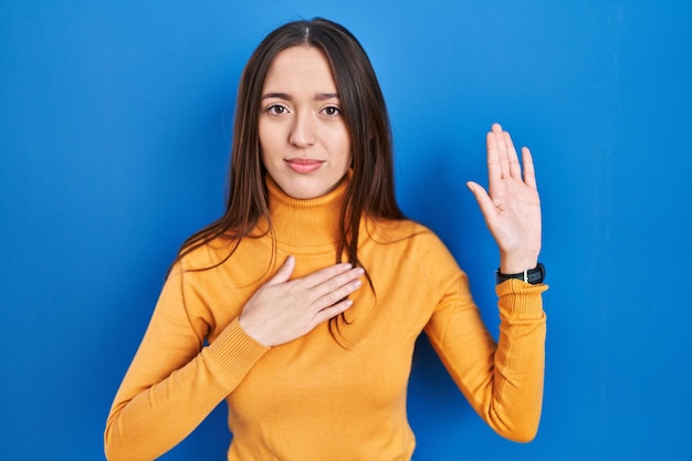 Young brunette woman standing over blue background swearing with hand on chest and open palm, making a loyalty promise oath