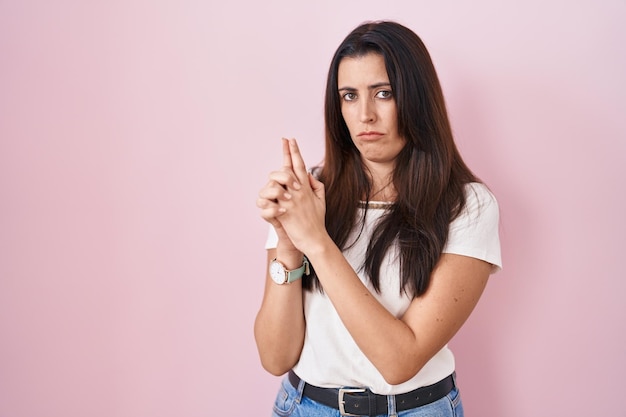 Young brunette woman standing over pink background holding symbolic gun with hand gesture, playing killing shooting weapons, angry face