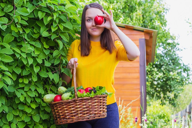 A young brunette woman stands in a bright yellow jacket in the garden with a wicker basket containing a crop of tomatoes, herbs and zucchini