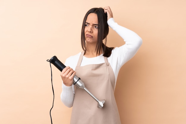 Young brunette woman using hand blender having doubts while scratching head