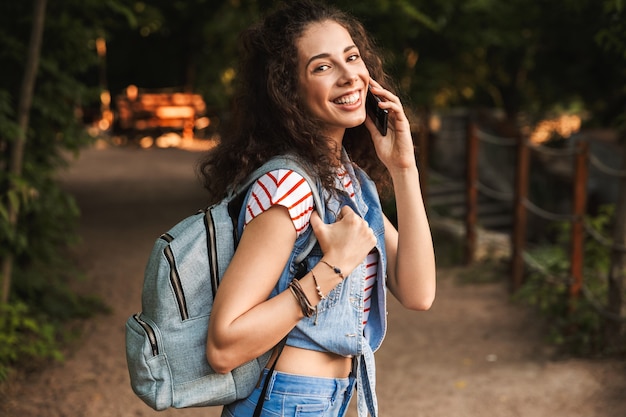 young brunette woman wearing backpack, smiling while walking through green park and speaking on mobile phone