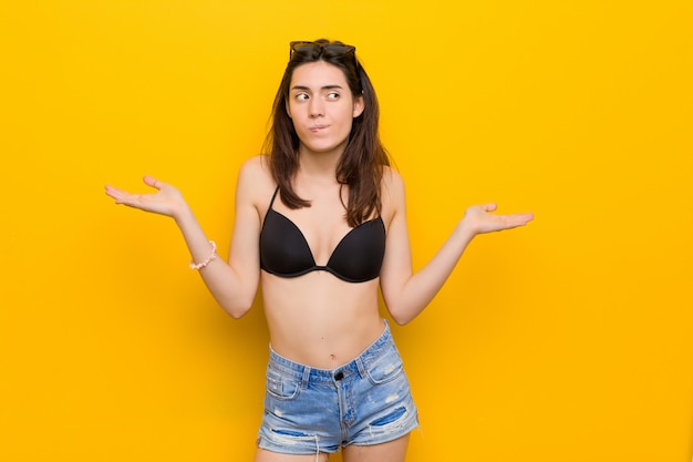 Young brunette woman wearing a bikini on yellow confused and doubtful shrugging shoulders to hold a copy space.