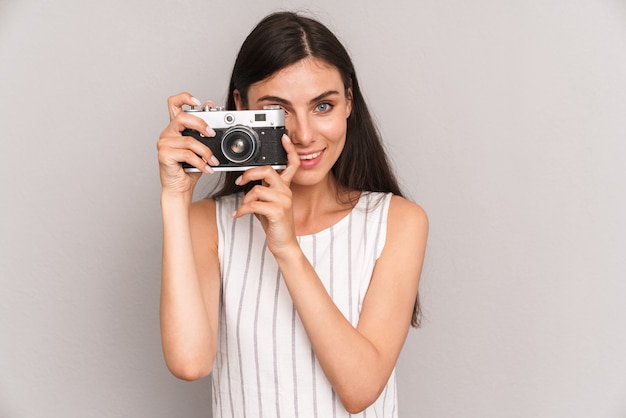  young brunette woman wearing dress smiling and taking photo on retro camera isolated over gray wall