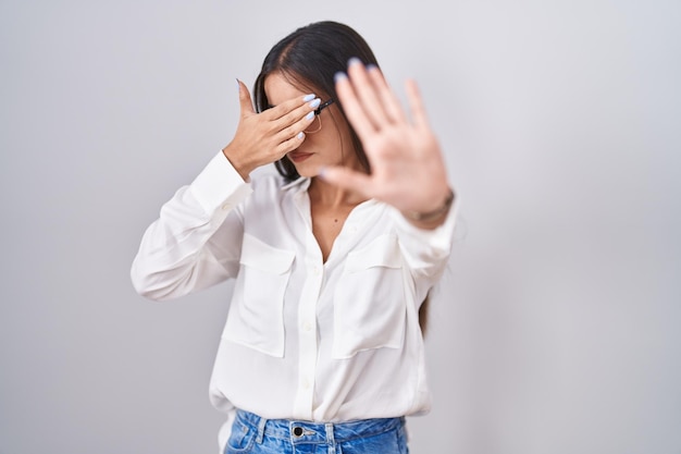 Young brunette woman wearing glasses covering eyes with hands and doing stop gesture with sad and fear expression embarrassed and negative concept