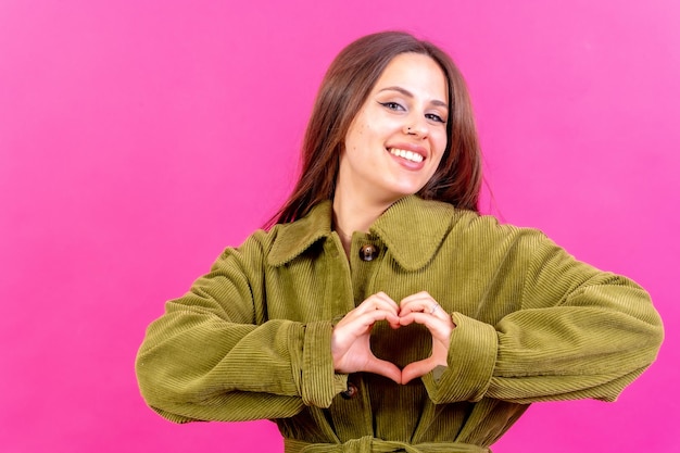 Young brunette woman wearing green sweater over isolated pink background smiling in love making heart symbol shape with hands romantic concept