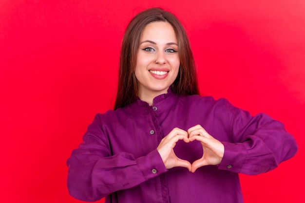 Young brunette woman wearing purple sweater over isolated red background smiling in love making heart symbol shape with hands romantic concept