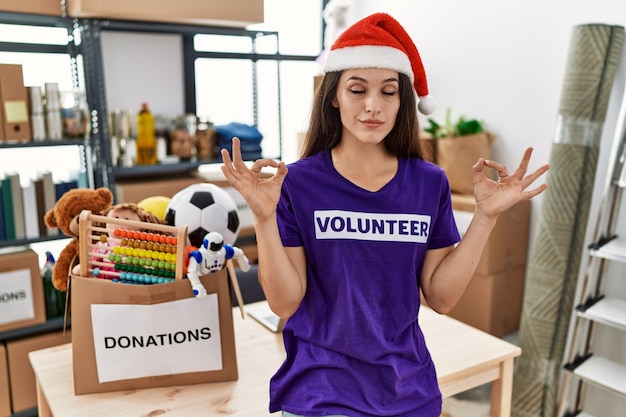 Young brunette woman wearing volunteer t shirt and christmas hat relax and smiling with eyes closed doing meditation gesture with fingers yoga concept