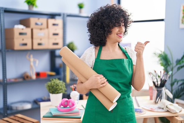 Young brunette woman with curly hair holding paper blueprints pointing thumb up to the side smiling happy with open mouth