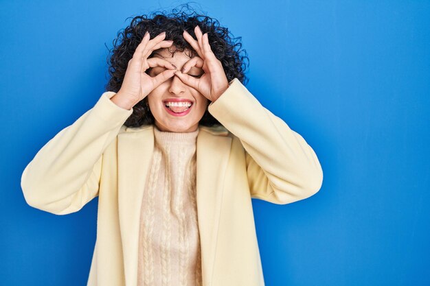 Young brunette woman with curly hair standing over blue background doing ok gesture like binoculars sticking tongue out eyes looking through fingers crazy expression