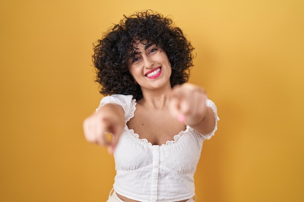 Young brunette woman with curly hair standing over yellow background pointing to you and the camera with fingers, smiling positive and cheerful