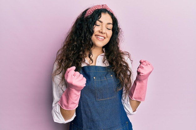 Young brunette woman with curly hair wearing cleaner apron and gloves excited for success with arms raised and eyes closed celebrating victory smiling. winner concept.