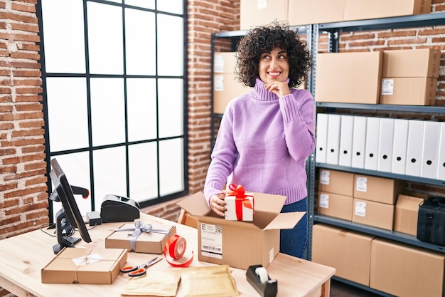 Young brunette woman with curly hair working at small business ecommerce preparing order serious face thinking about question with hand on chin thoughtful about confusing idea