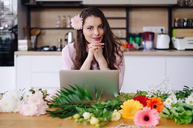 Young brunette woman with long wavy hair working on laptop with flowers on the desk with kitchen in background Talented florist developing online sales getting ready for workshop