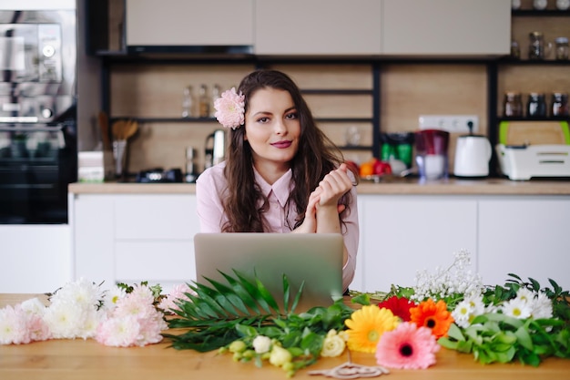 Young brunette woman with long wavy hair working on laptop with flowers on the desk with kitchen in background Talented florist developing online sales getting ready for workshop