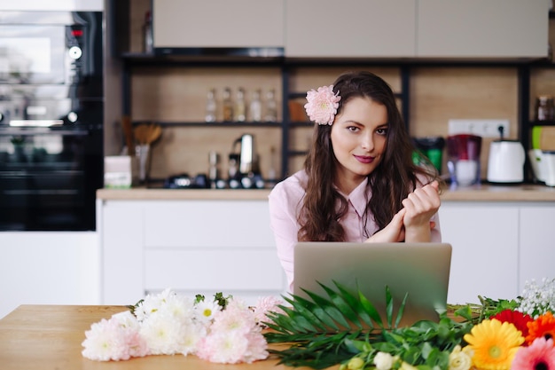 Young brunette woman with long wavy hair working on laptop with flowers on the desk with kitchen in background Talented florist developing online sales getting ready for workshop