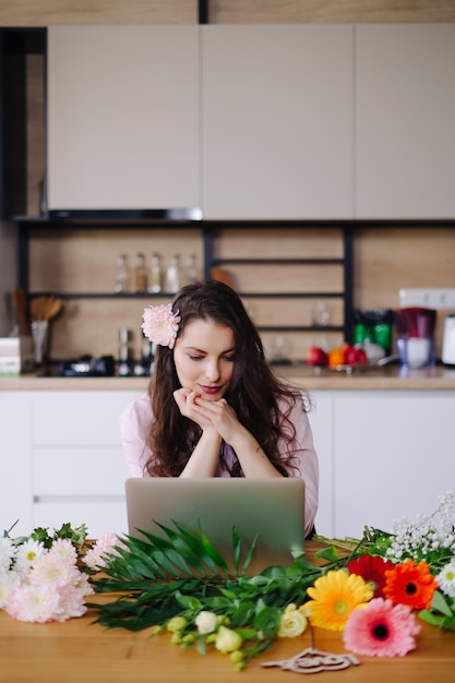 Young brunette woman with long wavy hair working on laptop with flowers on the desk with kitchen in background Talented florist developing online sales getting ready for workshop