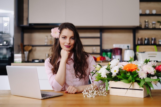 Young brunette woman with long wavy hair working on laptop with flowers on the desk with kitchen in background Talented florist developing online sales getting ready for workshop