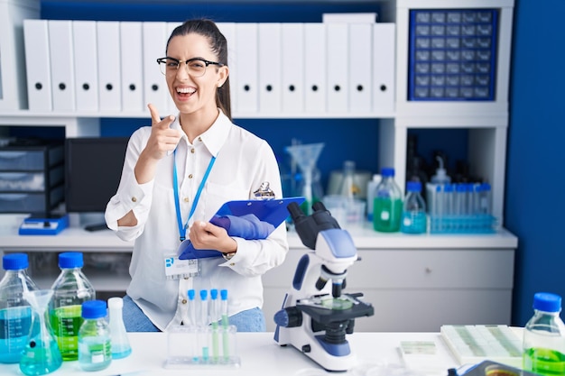 Young brunette woman working at scientist laboratory pointing fingers to camera with happy and funny face. good energy and vibes.