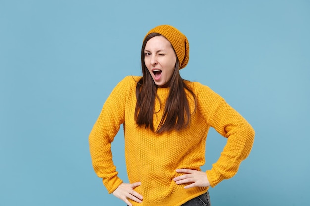 Young brunette woman in yellow sweater hat posing isolated on blue background studio portrait. People sincere emotions lifestyle concept. Mock up copy space. Stand with arms akimbo on waist blinking.