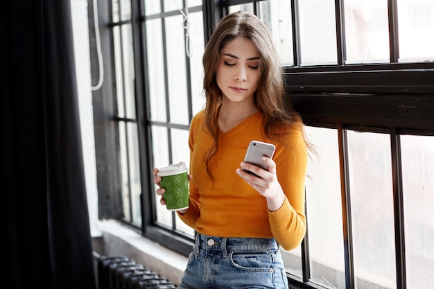 A young brunette in a yellow sweater stands by the window with a cup of coffee and a phone in her hand, talking on the phone