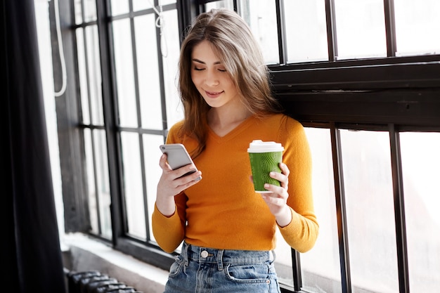 A young brunette in a yellow sweater stands by the window with a cup of coffee and a phone in her hand, talking on the phone