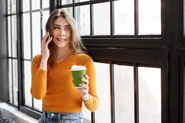 A young brunette in a yellow sweater stands by the window with a cup of coffee and a phone in her hand, talking on the phone