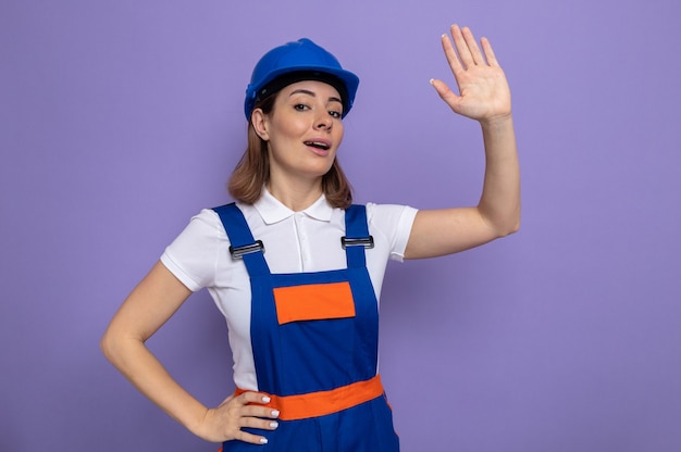 Young builder woman in construction uniform and safety helmet looking smiling confident waving with hand