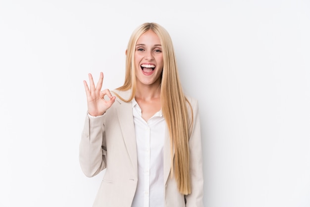 Young business blonde woman on white wall winks an eye and holds an okay gesture with hand.