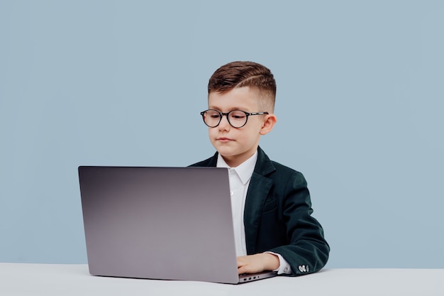 Young business boy with glasses using a laptop little boss in office funny kid isolated on blue back...