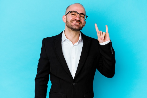 Young business caucasian bald man isolated on blue background showing a horns gesture as a revolution concept.