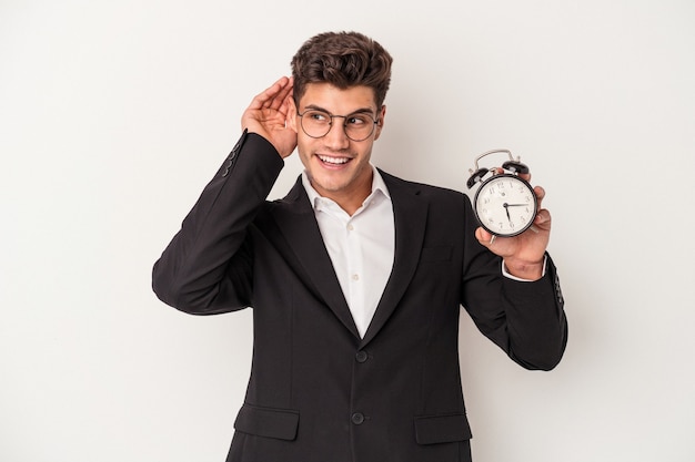 Young business caucasian man holding alarm clock isolated on white background trying to listening a gossip.