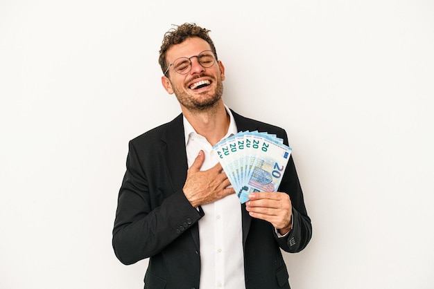 Young business caucasian man holding banknotes isolated on white background laughs out loudly keeping hand on chest.