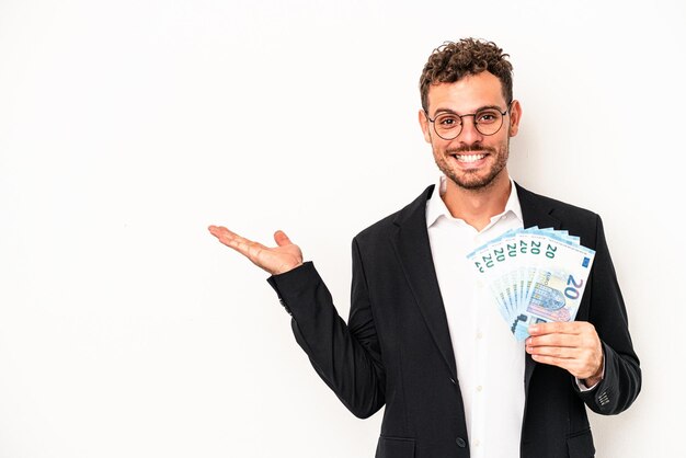 Young business caucasian man holding banknotes isolated on white background showing a copy space on a palm and holding another hand on waist.