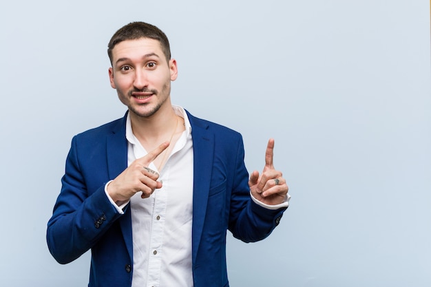 Young business caucasian man shocked pointing with index fingers to a copy space.