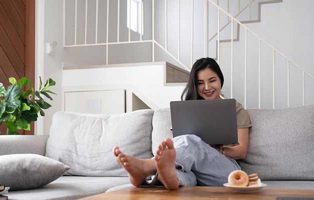 Young business freelance Asian woman working on laptop checking social media while lying on the sofa when relax in living room at home