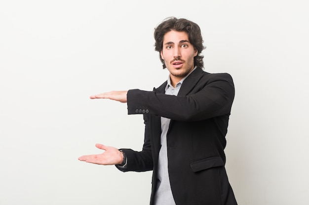 Young business man against a white wall shocked and amazed holding a blank space between hands.