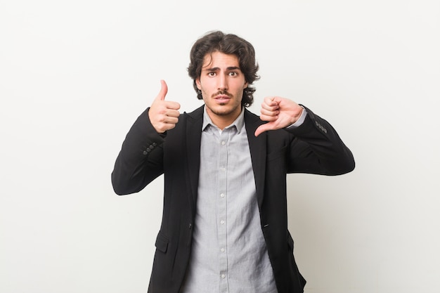 Young business man against a white wall showing thumbs up and thumbs down, difficult choose concept