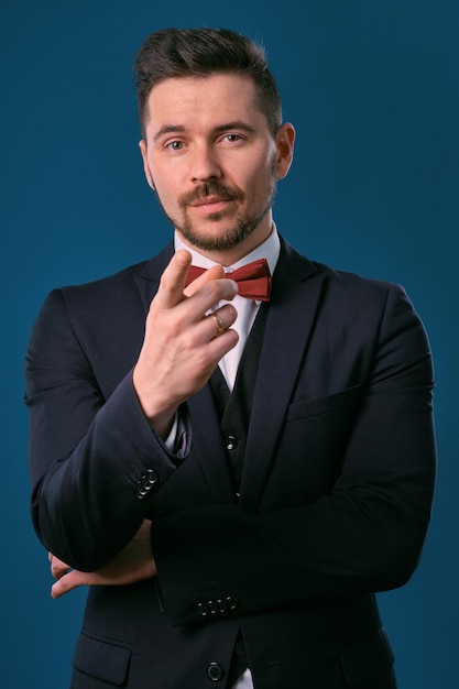 Young business man in classic black suit white shirt and red bowtie posing against a blue studio bac...