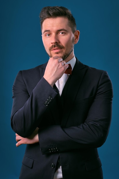 Young business man in classic black suit white shirt and red bowtie posing against a blue studio bac...