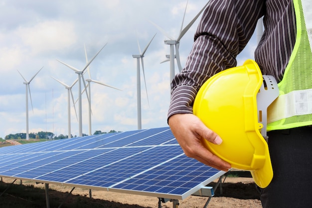 Young business man engineer hold yellow helmet at solar panel and wind generators power plant construction site background