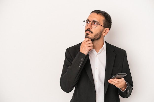 Young business man holding a mobile phone isolated on white background looking sideways with doubtful and skeptical expression.